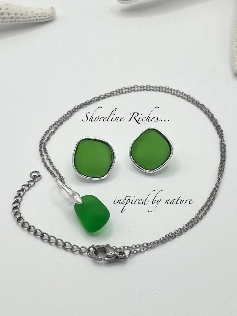 May include: A jewelry set featuring a necklace and matching earrings. The set includes a silver-toned chain necklace with a green pendant and a pair of green stud earrings. The text "Shoreline Riches... inspired by nature" is also visible.
