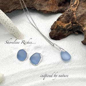 May include: A silver necklace and matching earrings set featuring light blue, teardrop-shaped pendants. The earrings and pendant are set in silver. The image includes the text "Shoreline Riches..." and "inspired by nature". The jewelry is displayed on a white, sandy surface.