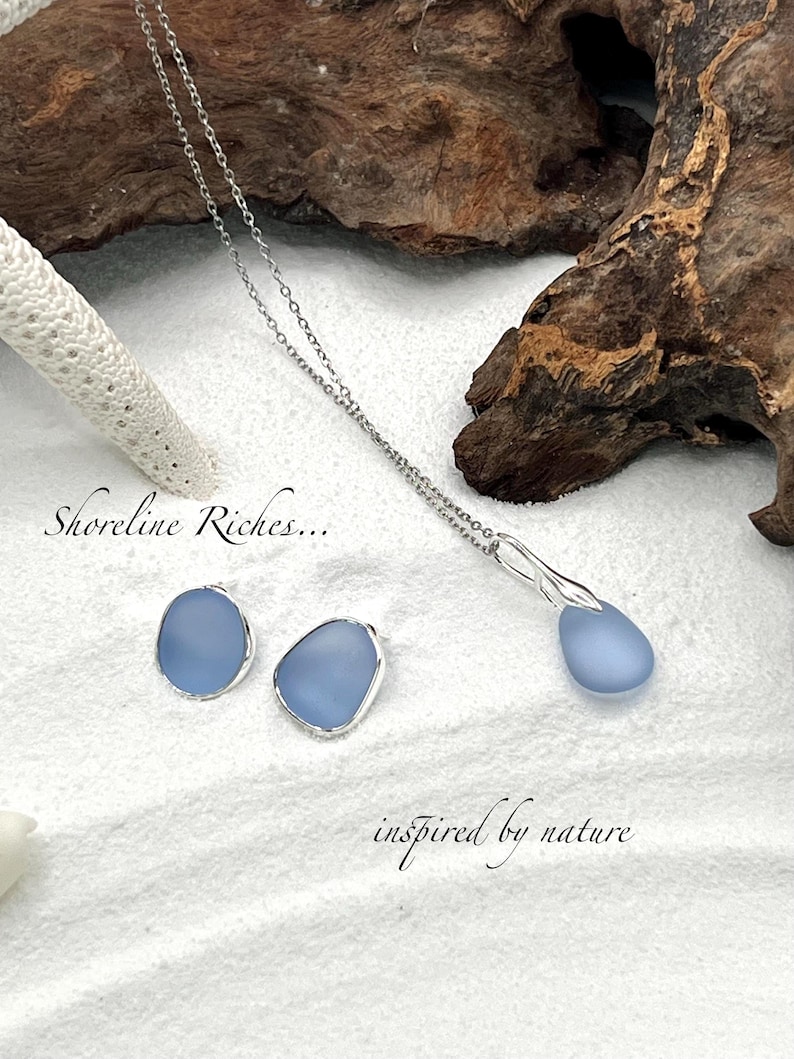 May include: A silver necklace and matching earrings set featuring light blue, teardrop-shaped pendants. The earrings and pendant are set in silver. The image includes the text "Shoreline Riches..." and "inspired by nature". The jewelry is displayed on a white, sandy surface.