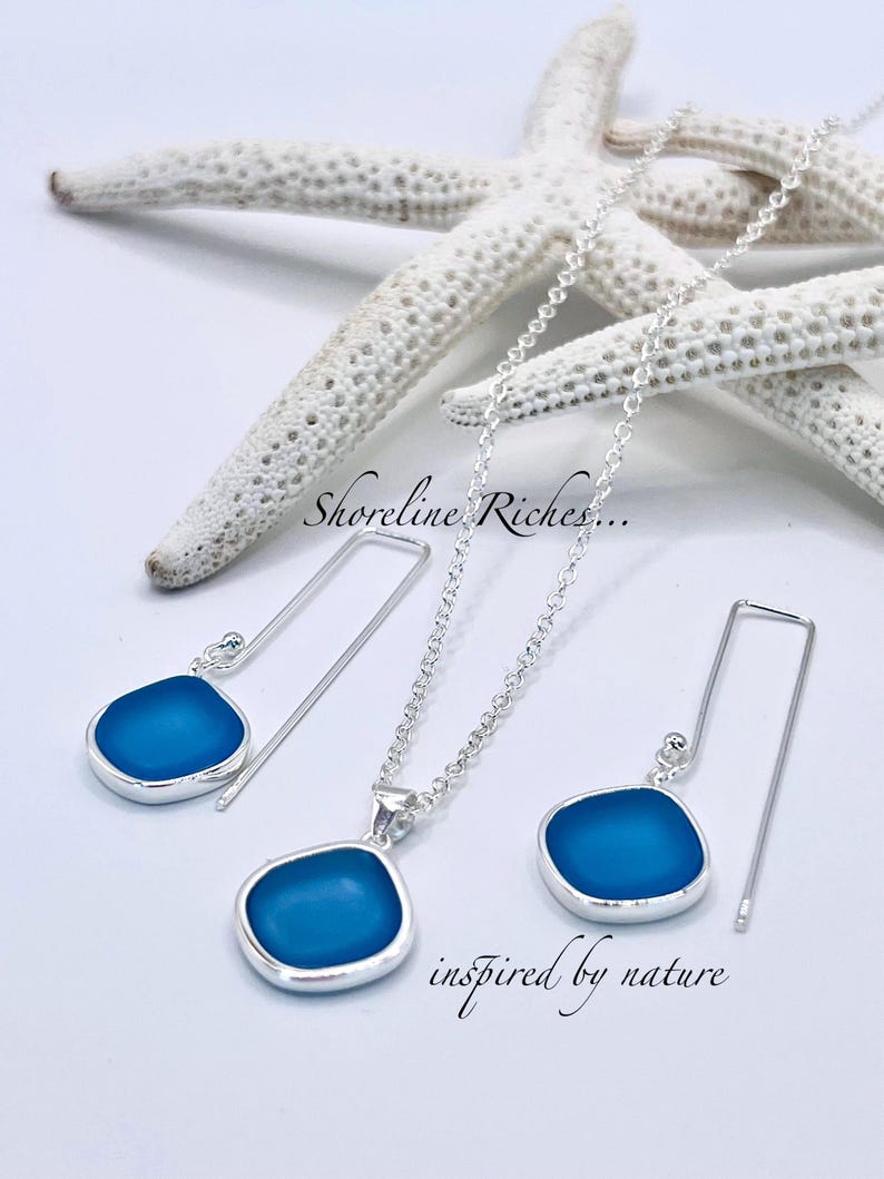 May include: A silver necklace and earrings set with a blue, sea glass-like pendant. The earrings have a long, thin hook design. The words "Shoreline Riches..." and "inspired by nature" are also visible.