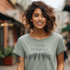 May include: A woman wearing a sage green t-shirt with a black graphic print that says "Forget the Village, I Need the Whole Vineyard, Mom Said".