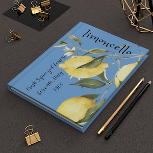 Puede incluir: Un diario azul con la palabra "limoncello" en escritura cursiva. La portada presenta una ilustración de acuarela de limones y hojas. El texto en la portada dice "Fresh Squeezed Lemons, Sorrento, Italy, 1900". Lápices y clips dorados y negros están cerca.