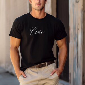 May include: Black crew neck t-shirt with the word "Ciao" in white script. The shirt is paired with beige trousers and a brown belt. The person is standing outside.