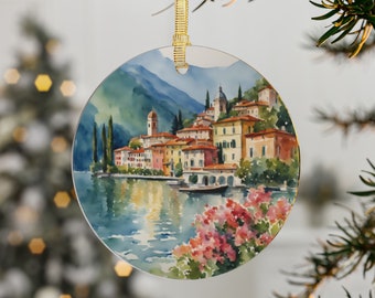 May include: A round, white, ceramic ornament with a watercolor painting of a small town on a lake. The town is surrounded by mountains and trees. The water is a light blue color and reflects the sky. There are pink flowers in the foreground.