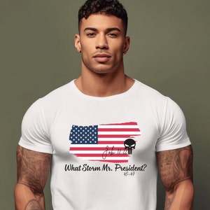 May include: White t-shirt with a red, white, and blue American flag graphic. The flag is painted in a brushstroke style. The text "What Storm Mr. President? 45-47" is printed below the flag. A black skull graphic is printed to the right of the flag.