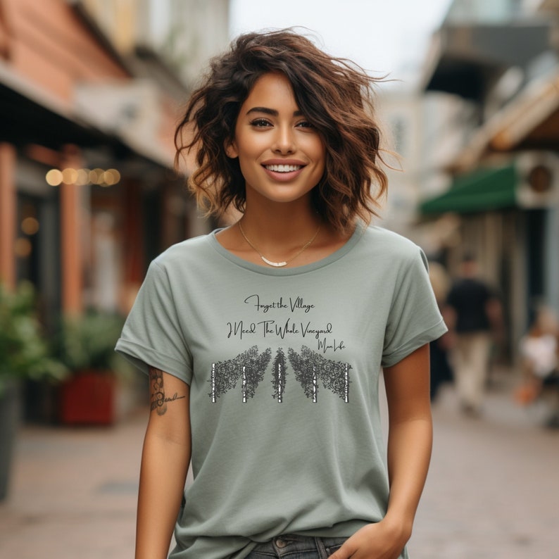 May include: A woman wearing a sage green t-shirt with a black graphic print that says "Forget the Village, I Need the Whole Vineyard, Mom Said".
