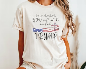 Könnte beinhalten: Ein beiges T-Shirt mit einem rot-weiß-blauen amerikanischen Flaggenmuster und einem Kreuz. Der Text "Be not deceived, GOD will not be mocked. Galatians 6:7" und "TRUMP" sind auf das T-Shirt gedruckt.