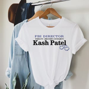 May include: White t-shirt with the text "FBI DIRECTOR" above "FIDELITY | BRAVERY | INTEGRITY" and "Kash Patel" in a bold font. A blue handcuff graphic is on the right. The shirt is hanging on a wooden hanger.