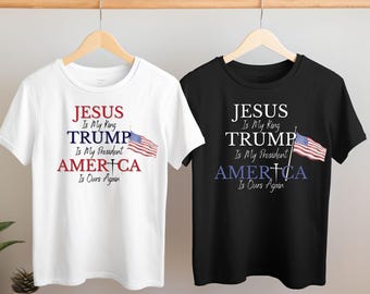 May include: Two t-shirts with a political slogan. The white t-shirt has a red, white, and blue American flag graphic. The black t-shirt has a blue American flag graphic. Both t-shirts have the text "JESUS Is My King TRUMP Is My President AMERICA Is Ours Again".