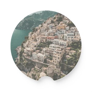 May include: An aerial view of a coastal town with white buildings clustered together on a hillside. The town is surrounded by lush green vegetation and a blue body of water.