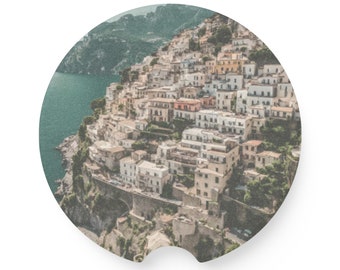 May include: An aerial view of a coastal town with white buildings clustered together on a hillside. The town is surrounded by lush green vegetation and a blue body of water.