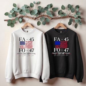 May include: Two sweatshirts with a distressed American flag design. The white sweatshirt has a blue and red flag with the text "FA-45 FO-47 TRUMP We The People One Nation Under God". The black sweatshirt has a blue and red flag with the text "FA-45 FO-47 TRUMP We The People One Nation Under God".