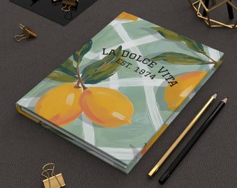 May include: A hardcover journal with a lemon and leaf design. The cover features the text "LA DOLCE VITA EST. 1974" over a green and white grid pattern. Two pencils and gold binder clips are also visible.