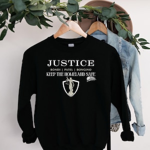 May include: Black sweatshirt with white text that reads "JUSTICE BONDI | PATEL | BONGINO KEEP THE HOMELAND SAFE". The text is centered on the sweatshirt and is surrounded by a white shield with a figure of justice holding scales and a sword.