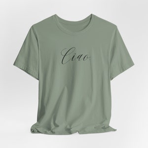 May include: A sage green t-shirt featuring the word "Ciao" in black cursive script. This casual, short-sleeved top is made from a soft fabric. "Ciao" is an Italian greeting, suitable for everyday wear.