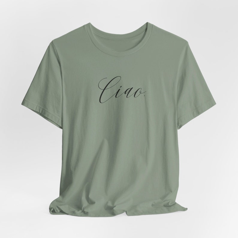 May include: A sage green t-shirt featuring the word "Ciao" in black cursive script. This casual, short-sleeved top is made from a soft fabric. "Ciao" is an Italian greeting, suitable for everyday wear.