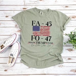 May include: A green t-shirt with a distressed American flag design and the text "FA-45 FO-47 TRUMP America First We The People One Nation Under God".