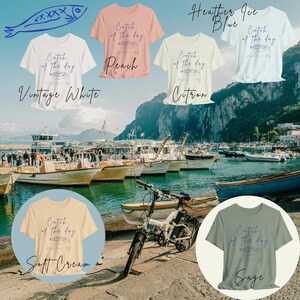 May include: A collection of ten t-shirts in various colors, each featuring a graphic of a fish with the text "Catch of the day". The t-shirts are displayed on a background of a harbor with boats and water.