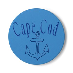 May include: A round, blue coaster with the words "Cape Cod" in a script font and a navy blue anchor design. The coaster is a solid blue color and has a slightly textured surface. The anchor is centered below the text.