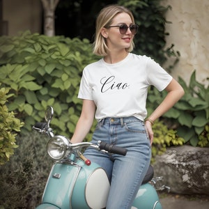 May include: A woman wearing a white t-shirt with the word "Ciao" in black script, blue jeans, and sunglasses, sitting on a light blue scooter. The scooter has a white seat and chrome accents. Green foliage is in the background.