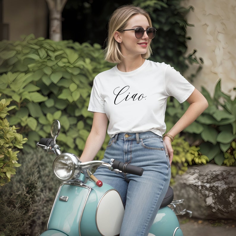 May include: A woman wearing a white t-shirt with the word "Ciao" in black script, blue jeans, and sunglasses, sitting on a light blue scooter. The scooter has a white seat and chrome accents. Green foliage is in the background.