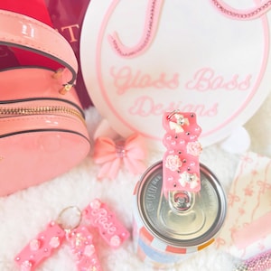 Puede incluir: Accesorios rosas, incluyendo un pequeño bolso, un llavero con el nombre "Rose" y un abre-latas decorado con flores y pedrería. Un letrero blanco con el texto "Glass Boss Designs" se encuentra en el fondo.