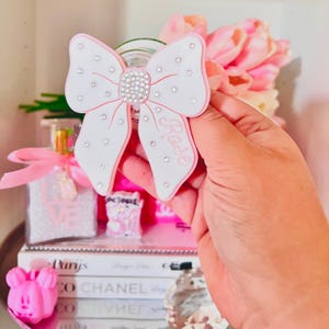 Puede incluir: Un accesorio en forma de lazo blanco y rosa con el nombre "Rose" escrito. El lazo está adornado con pedrería y sostenido por una mano. El fondo incluye un frasco de perfume, libros y flores rosas.