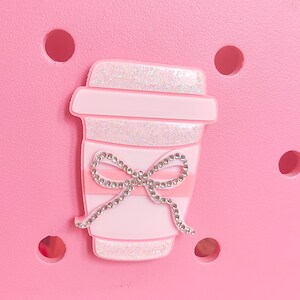 Op de afbeelding: Een roze schoenbedel in de vorm van een koffiekopje, versierd met een sprankelende strik. De beker heeft glitterdetails aan de boven- en onderkant, met een roze band. De bedel staat op een roze achtergrond met verschillende ronde gaten.