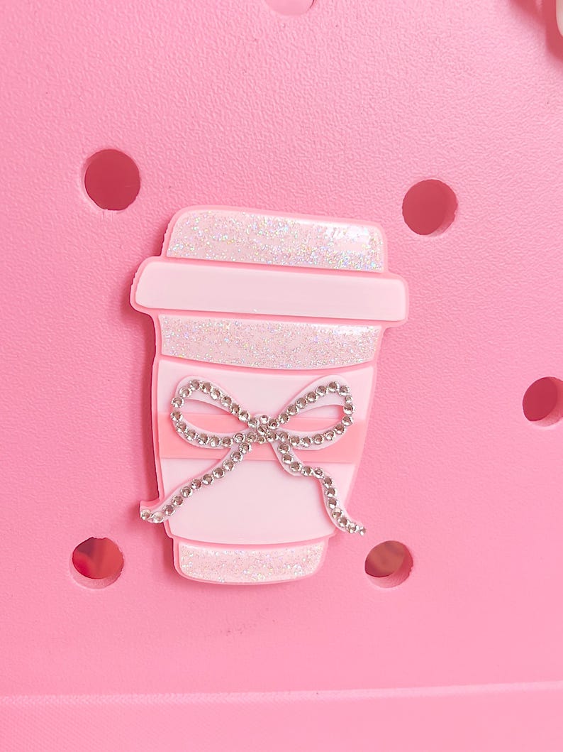 Op de afbeelding: Een roze schoenbedel in de vorm van een koffiekopje, versierd met een sprankelende strik. De beker heeft glitterdetails aan de boven- en onderkant, met een roze band. De bedel staat op een roze achtergrond met verschillende ronde gaten.