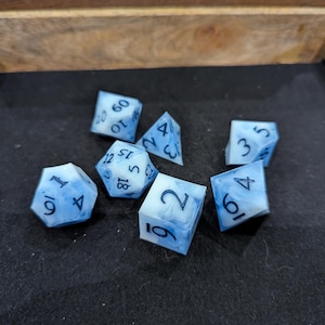 May include: Set of seven polyhedral dice with a blue and white marbled design. The dice are arranged on a black surface inside a wooden tray. Each die has white numbers.
