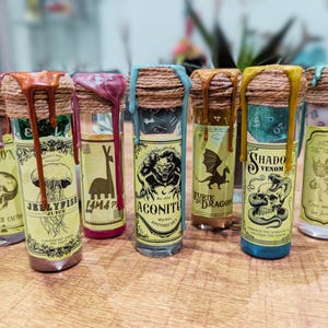 Puede incluir: Una colección de botellas de pociones de vidrio con tapones de corcho y goteos de cera coloridos. Cada botella tiene una etiqueta única con ilustraciones y texto, incluyendo una calavera, una medusa y un dragón. Las botellas están dispuestas sobre una superficie de madera.