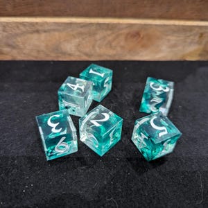 Puede incluir: Seis dados de juego de color verde azulado y transparentes con números plateados están esparcidos sobre una superficie negra. Los dados son translúcidos, lo que permite vislumbrar el diseño interior. Los dados están dentro de una bandeja de madera.