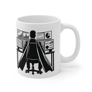 Puede incluir: Taza de café de cerámica blanca con una ilustración en blanco y negro de una persona con capa de superhéroe sentada en un escritorio con múltiples monitores de computadora que muestran gráficos. La taza tiene un asa curva.