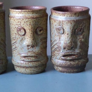 May include: A set of six cylindrical ceramic mugs, each featuring a hand-sculpted face with distinct eyes, a nose, and a mouth. The mugs have a speckled, tan-colored glaze with a reddish-brown interior rim and a textured, earthy appearance.