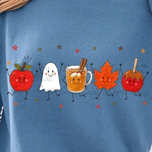 May include: A blue long-sleeve sweatshirt featuring a graphic print of five cartoon characters: a red apple, a white ghost, a mug of cider with cinnamon sticks, an orange maple leaf, and a red candy apple, all accented by small colorful stars.