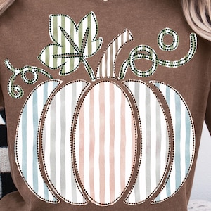Könnte beinhalten: Braunes T-Shirt mit einem farbenfrohen Kürbis-Design. Der Kürbis hat vertikale Streifen in Rosa, Blau und Grau. Ein grünes Blatt und Ranken ergänzen das herbstliche Grafikdesign.