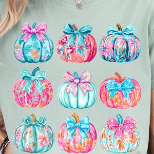 May include: A light green t-shirt featuring nine colorful pumpkin illustrations. Each pumpkin has a different floral pattern and is topped with a decorative bow. The pumpkins are in shades of pink, blue, orange, and teal.