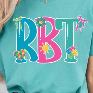 May include: A teal t-shirt featuring the letters "RBT" in blue, pink, and teal. The design includes a heart, flower, sun, and puzzle pieces, all in vibrant colors. The shirt has a round neckline.