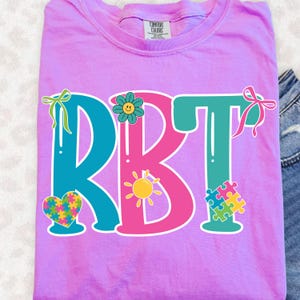 May include: A lilac-colored t-shirt with the letters "RBT" in teal and pink, decorated with a flower, sun, heart, and puzzle pieces. The shirt has a crew neck and short sleeves.
