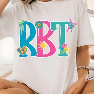 May include: White t-shirt with the letters "RBT" in teal and pink, decorated with a heart, flower, sun, and puzzle pieces. The design includes bows and a smiling flower. The shirt is casual and comfortable.