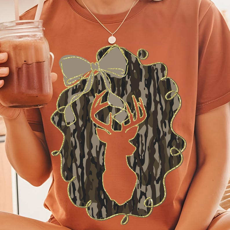Puede incluir: Camiseta de color &oacute;xido con una silueta de ciervo dentro de un dise&ntilde;o de corona de camuflaje. Un lazo gris con detalles de purpurina dorada se encuentra encima del ciervo. El dise&ntilde;o est&aacute; delineado con purpurina dorada.