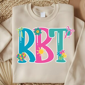 May include: A beige sweatshirt with the letters "RBT" in blue, pink, and teal. The design includes a heart-shaped puzzle, a sun, a flower, and puzzle pieces. The letters are outlined in white.