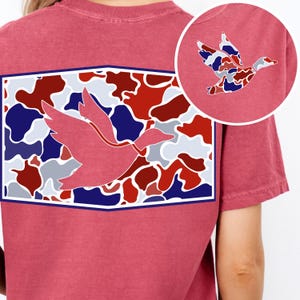 K&ouml;nnte beinhalten: Staubrosa T-Shirt mit einer gro&szlig;en Grafik einer fliegenden Ente. Die Ente ist rosa umrandet und vor einem Camouflagemuster aus Rot, Wei&szlig; und Blau platziert. Eine kleinere, kreisf&ouml;rmige Grafik der Ente ist ebenfalls sichtbar.