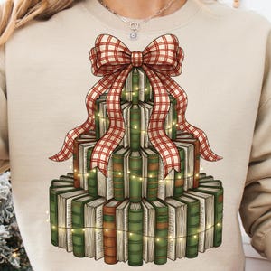 May include: A cream-colored sweatshirt featuring a tiered stack of books designed to resemble a Christmas tree. A large red and white plaid bow sits atop the books, with string lights wrapped around the books. The books are green and brown.