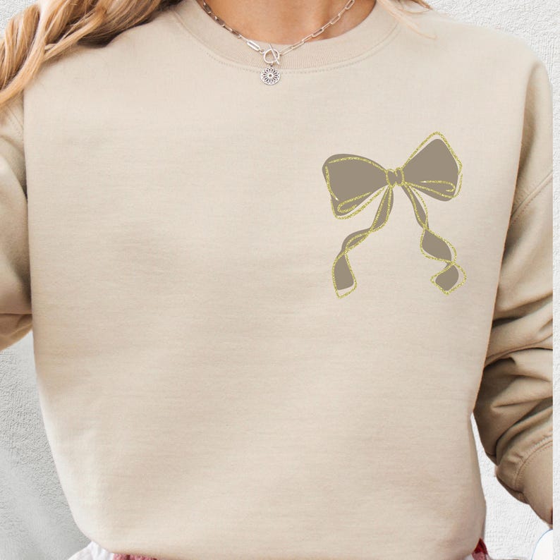 Puede incluir: Sudadera beige con un gran dise&ntilde;o de lazo en la parte delantera. El lazo es de color gris apagado con contornos dorados brillantes. La sudadera tiene mangas largas y cuello redondo.
