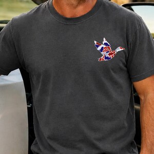 K&ouml;nnte beinhalten: Dunkelgraues T-Shirt mit einem bunten Entenmuster auf der Brust. Das Entenmuster besteht aus roten, wei&szlig;en und blauen Mustern. Das T-Shirt hat einen Rundhalsausschnitt und kurze &Auml;rmel.