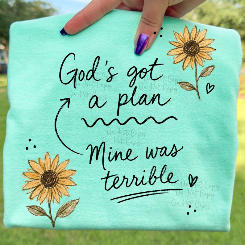 Puede incluir: Una camiseta verde menta con un estampado gr&aacute;fico negro que dice God's got a plan, con una flecha curva que apunta al texto, seguida de las palabras Mine was terrible. Dos girasoles amarillos con centros marrones aparecen en la prenda.