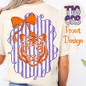 May include: Cream-coloured t-shirt with a tiger design. The back features an orange bow, a tiger head, and purple and white stripes. The front design says "Tigers" in purple and orange.