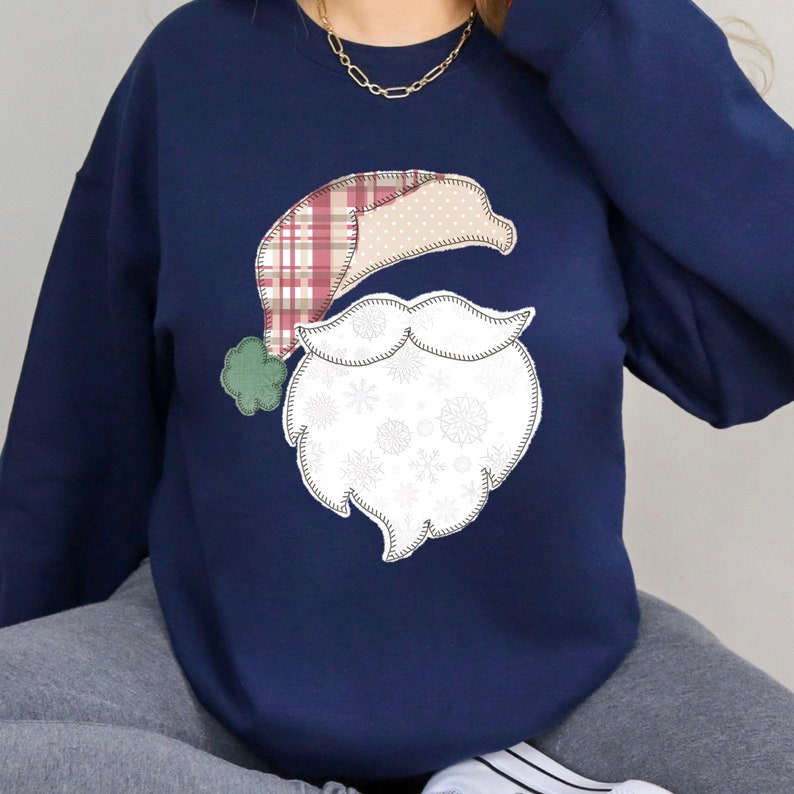 May include: A navy blue sweatshirt with a white and red patchwork Santa Claus design. The Santa Claus design has a white beard with snowflakes and a red and white plaid hat.