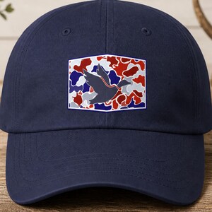 K&ouml;nnte beinhalten: Marineblaue Baseballkappe mit einem rechteckigen Aufn&auml;her. Der Aufn&auml;her zeigt eine Entensilhouette vor einem Tarnmuster in Rot, Wei&szlig; und Blau. Die Kappe ist aus weichem Material und hat einen gebogenen Schirm.